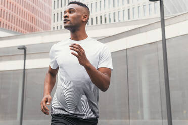 From below of fit black male athlete in sportswear running while working out in downtown of modern city in morning and looking away - ADSF45929