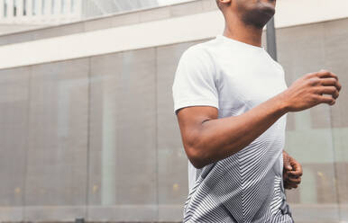 Side view of anonymous crop fit black male athlete in sportswear running while working out in downtown of modern city in morning - ADSF45930