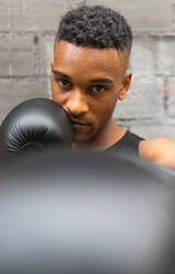 Portrait of African American male boxer in sportswear and leather gloves practicing punch and looking at camera against brick wall in gym - ADSF45931