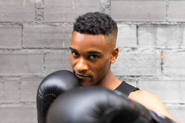 Portrait of African American male boxer in sportswear and leather gloves practicing punch and looking at camera against brick wall in gym - ADSF45932