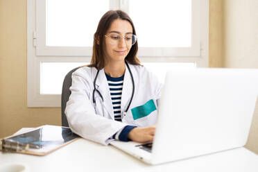 Young female doctor in medical uniform and glasses sitting at table with laptop and giving online consultation in office - ADSF45933