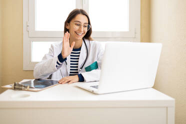Positive young female doctor in white cloak waving hand to patient while having video call on laptop in office - ADSF45936