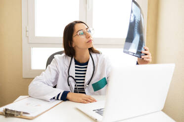 Positive young female doctor in white cloak and glasses reading x ray while sitting at table in office - ADSF45937