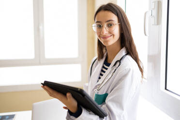 Young female doctor in white coat and eyeglasses looking at camera of tablet while standing in office of modern clinic - ADSF45940