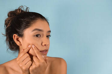 Young focused Hispanic female with bare shoulders looking away while squeezing pimple on face in studio - ADSF45941