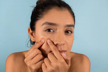 Young focused Hispanic female with bare shoulders looking at camera while squeezing pimple on face in studio - ADSF45943