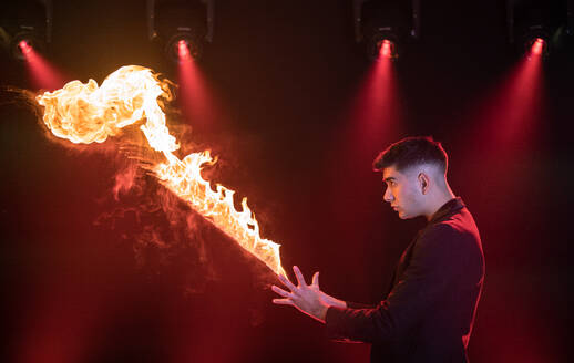 Seitenansicht eines männlichen Zauberkünstlers in schwarzem Anzug, der einen Zaubertrick mit brennender Flamme auf einer dunklen Zirkusbühne vorführt - ADSF45947