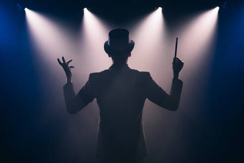 Back view of male illusionist in suit and top hat with magic stick making presentation on dark stage in circus - ADSF45948