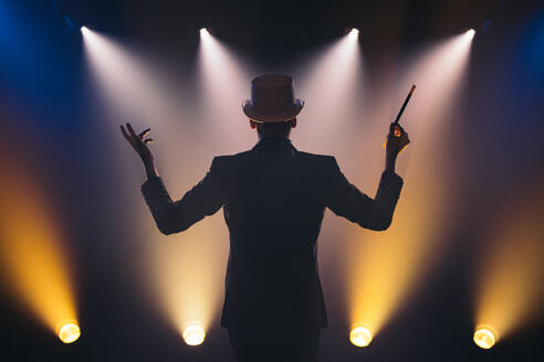 Back view of male illusionist in suit and top hat with magic stick making presentation on dark stage in circus - ADSF45949