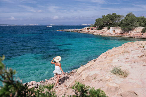 Back view of unrecognizable female tourist in hat walking on rocky cliff and enjoying summer vacation near turquoise sea in Balearic Islands - ADSF45954