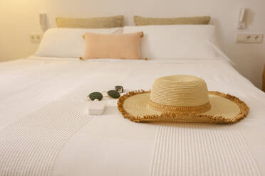 Sunhat and sunglasses on bed in hotel with white and beige pillows and blanket on summer vacation in Balearic Islands - ADSF45955