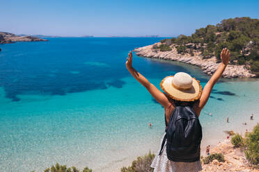 Back view of anonymous female tourist in hat celebrating summer holiday with raised arms and admiring amazing view of blue sea on Balearic Islands - ADSF45958
