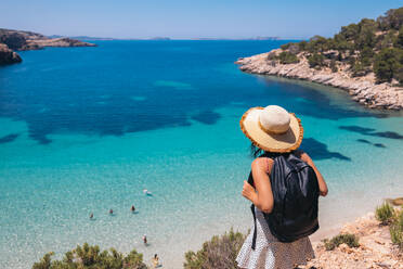 Back view of anonymous female tourist in hat with backpack admiring amazing view of blue sea on Balearic Islands - ADSF45959