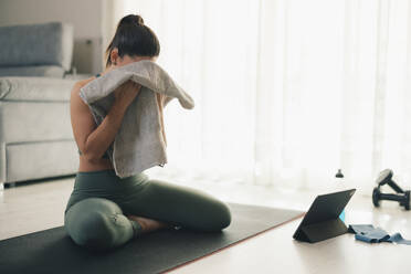 Side view of unrecognizable young female in sportswear sitting on mat on floor with tablet, dumbbells and wiping face with towel while taking break from yoga exercise in living room - ADSF45961