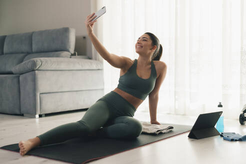 Sportliche Frau macht eine Pause vom Yoga, um ein Selfie zu machen und ihr Telefon und Tablet zu Hause zu überprüfen - ADSF45963