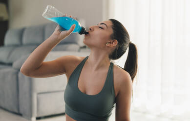 Young barefoot female in sportswear with closed eyes while drinking water from bottle while sitting and taking break in yoga practice at home - ADSF45964