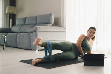 Side view of slim barefoot smiling young female lying on mat in plank position and looking at screen of tablet while attending online session and stretching legs with yoga bands in living room - ADSF45966