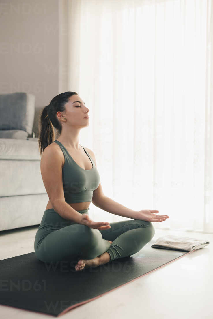 Full body side view of fit female in sportswear sitting on mat in lotus  position and meditating while practicing yoga at home stock photo