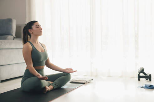 Full body side view of fit female in sportswear sitting on mat in lotus position and meditating while practicing yoga at home - ADSF45969