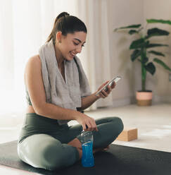 Smiling young female in sportswear with towel around neck using smartphone while sitting with bottle of energy drink on yoga mat at home - ADSF45973