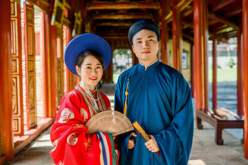 Positive Asian couple in gorgeous and colorful traditional clothes with headwear standing on blurred background of building passage in Hue Vietnam and looking at camera in daylight - ADSF45988