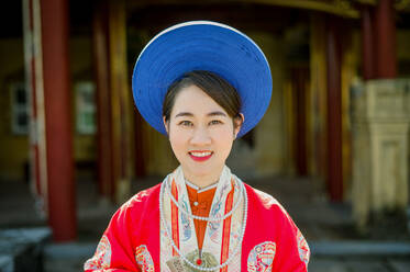 Portrait of positive young Asian female in traditional outfit with hat and makeup looking at camera while standing on against blurred apartment background in Hue Vietnam - ADSF45989