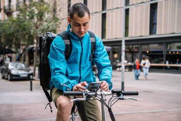 Delivery man in bright blue jacket with backpack sitting on bicycle in city street and using phone while working in Barcelona - ADSF45995