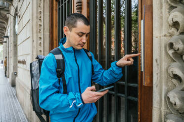 Side view of man in blue service jacket and with delivery backpack using phone while calling on intercom to customer - ADSF45996