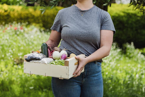 Crop anonymous young female in casual clothes carrying boxful of various fresh organic vegetables and seeds in covers while standing in agriculture field with blurred blooming flower plants in daylight - ADSF46011