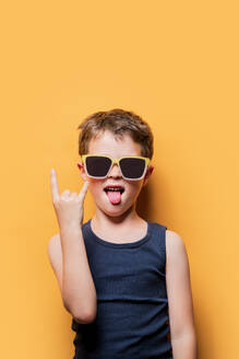 Preteen kid in tank top and sunglasses showing rock and roll sign on yellow background - ADSF46016