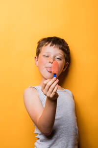 Cheerful boy in gray t shirt with tongue out and eye closed showing fruit ice pop against orange background - ADSF46018