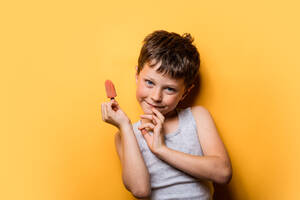 Smiling boy in gray t shirt with fruit ice pop looking at camera against orange background - ADSF46019