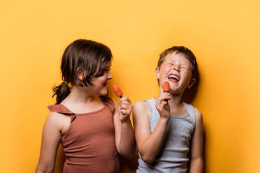 Funny little boy laughing brightly while standing in studio with girl eating yummy sweet ice pops against orange background having fun together - ADSF46021