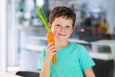 Portrait of positive cute boy in casual clothes smiling and looking at camera while standing at kitchen table with carrot against blurred background - ADSF46023
