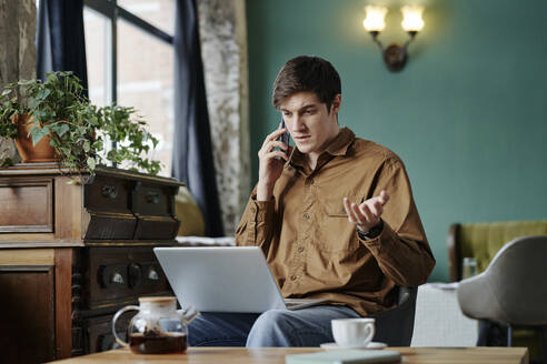 Geschäftsmann, der mit seinem Laptop in einem Café sitzt und mit seinem Smartphone spricht - DSHF00902
