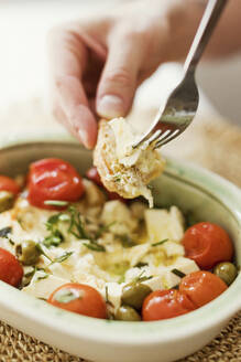 Hand einer Person, die gebackenen Feta-Käse mit Oliven, Kirschtomaten und Kräutern isst - ONAF00612