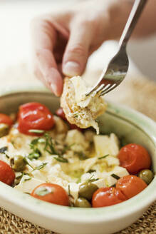 Hand of person eating baked feta cheese with olives, cherry tomatoes and herbs - ONAF00612