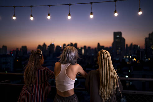 Friends standing under string lights on rooftop at dusk - IKF01016