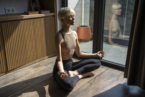 Blond woman with eyes closed meditating on floor at home - YBF00028