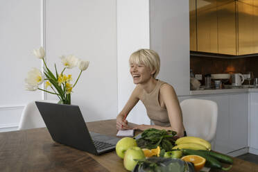 Lächelnde Ernährungsberaterin, die zu Hause am Laptop Notizen zur Ernährung macht - YBF00034