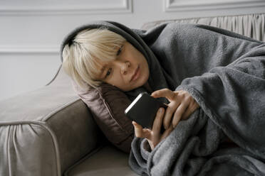 Blond woman using smart phone lying on sofa at home - YBF00055