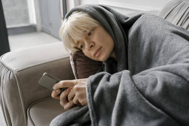 Blond woman using mobile phone lying on sofa at home - YBF00056