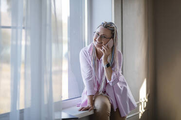 Lächelnde Frau, die zu Hause am Fenster sitzt und mit ihrem Smartphone telefoniert - ADF00128