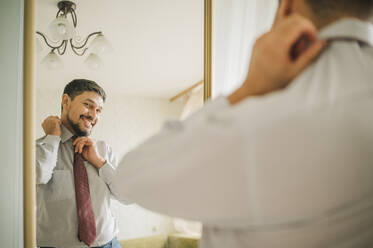 Reflection of smiling man tying necktie looking in mirror at home - ANAF01866