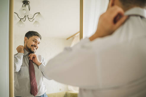 Reflektion eines lächelnden Mannes, der eine Krawatte bindet, im Spiegel zu Hause - ANAF01866