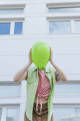 Frau bedeckt Gesicht mit grünem Luftballon vor einem Gebäude - MGRF01040