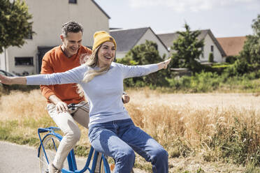 Cheerful man riding bicycle with woman in front of houses - UUF29660