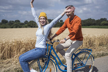 Cheerful woman with arms raised enjoying bicycle ride with man on sunny day - UUF29666