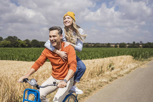 Glückliches Liebespaar, das an einem sonnigen Tag vor einem Feld Fahrrad fährt - UUF29677