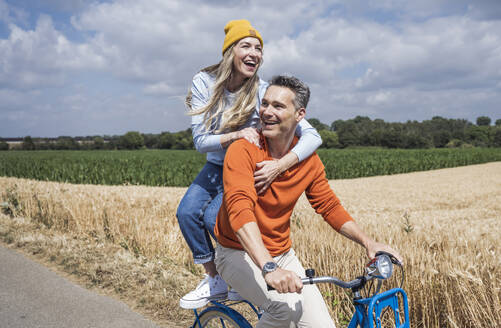 Fröhliches, liebendes Paar, das an einem sonnigen Tag vor einem Feld Fahrrad fährt - UUF29678